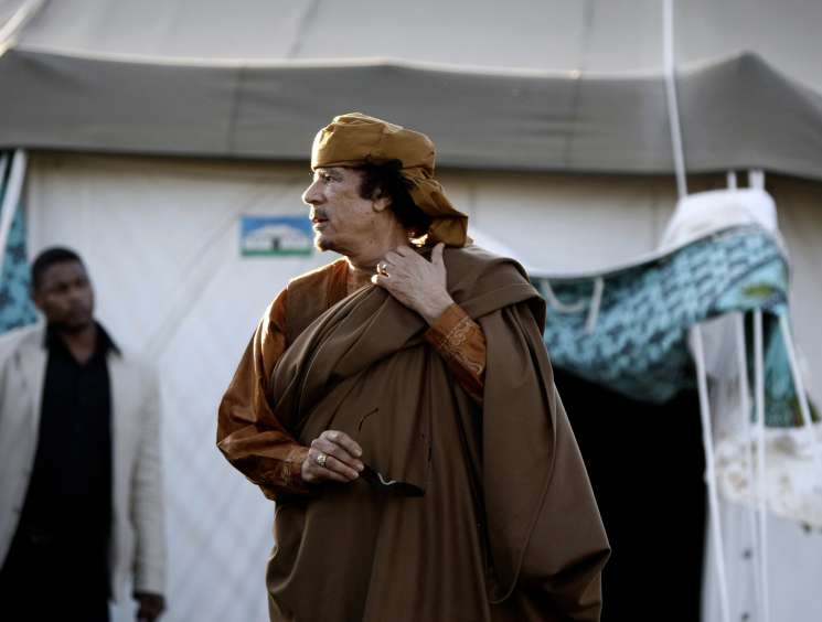 Man in brown traditional clothing and cap standing outside a tent structure.