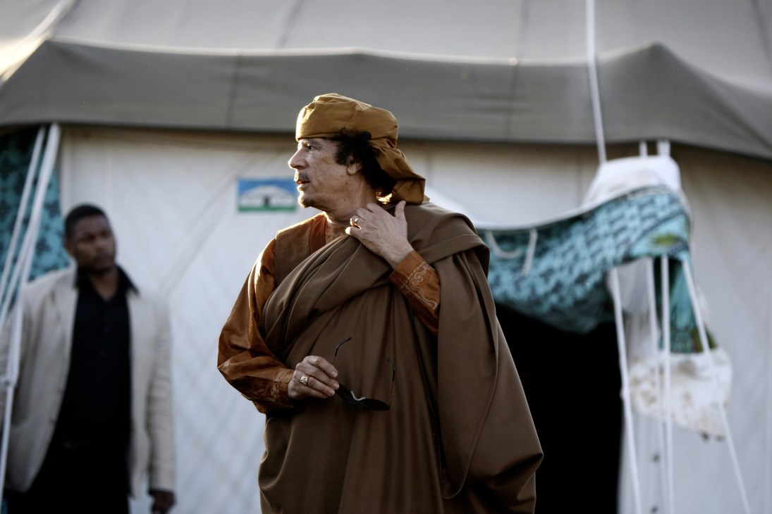Man in brown traditional clothing and cap standing outside a tent structure.