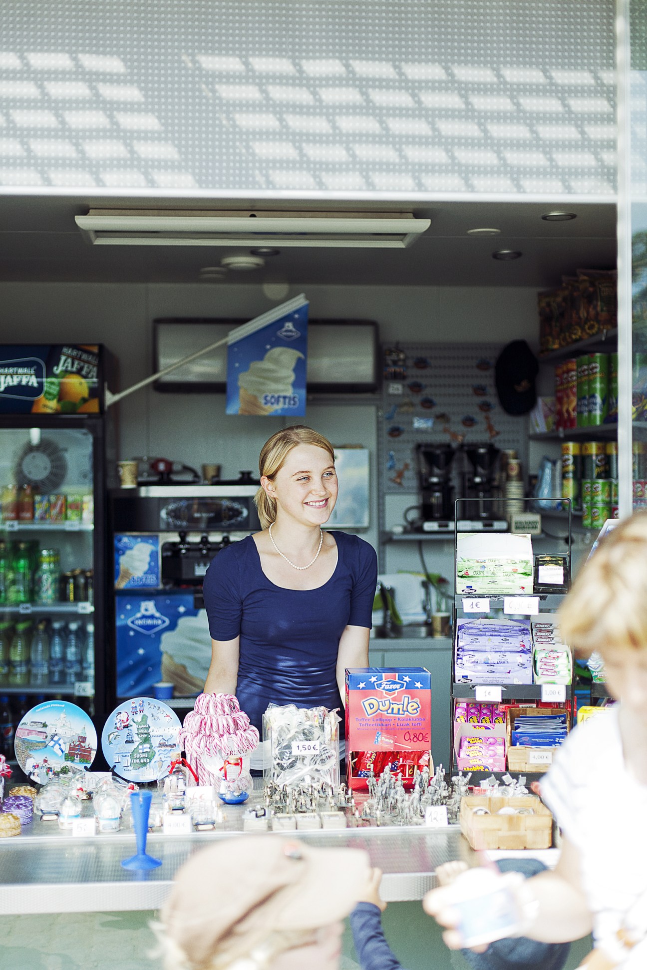 A smiling young woman working behind the counter of an ice cream kiosk, with shelves of products visible behind her.