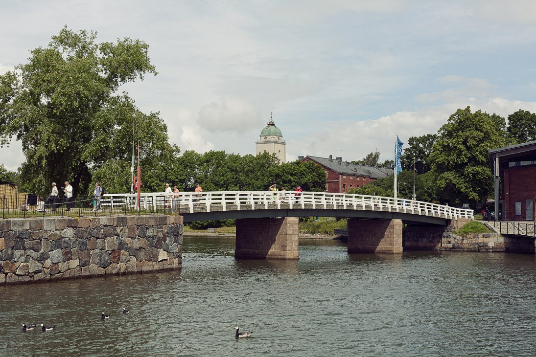 White pedestrian bridge spanning water between two islands with stone fortress walls, historic buildings and a domed tower visible beyond.