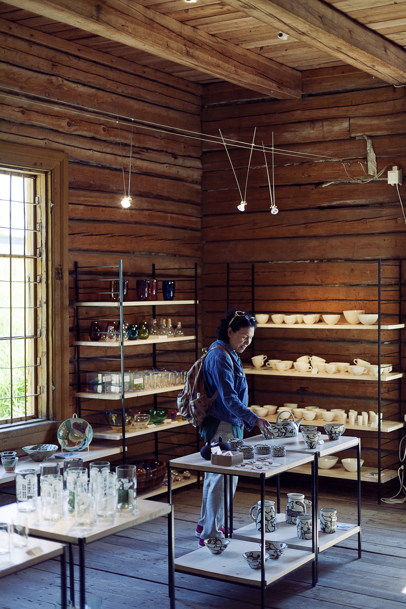 Woman in blue shirt standing in wooden cabin displaying ceramic vessels on shelves and tables.