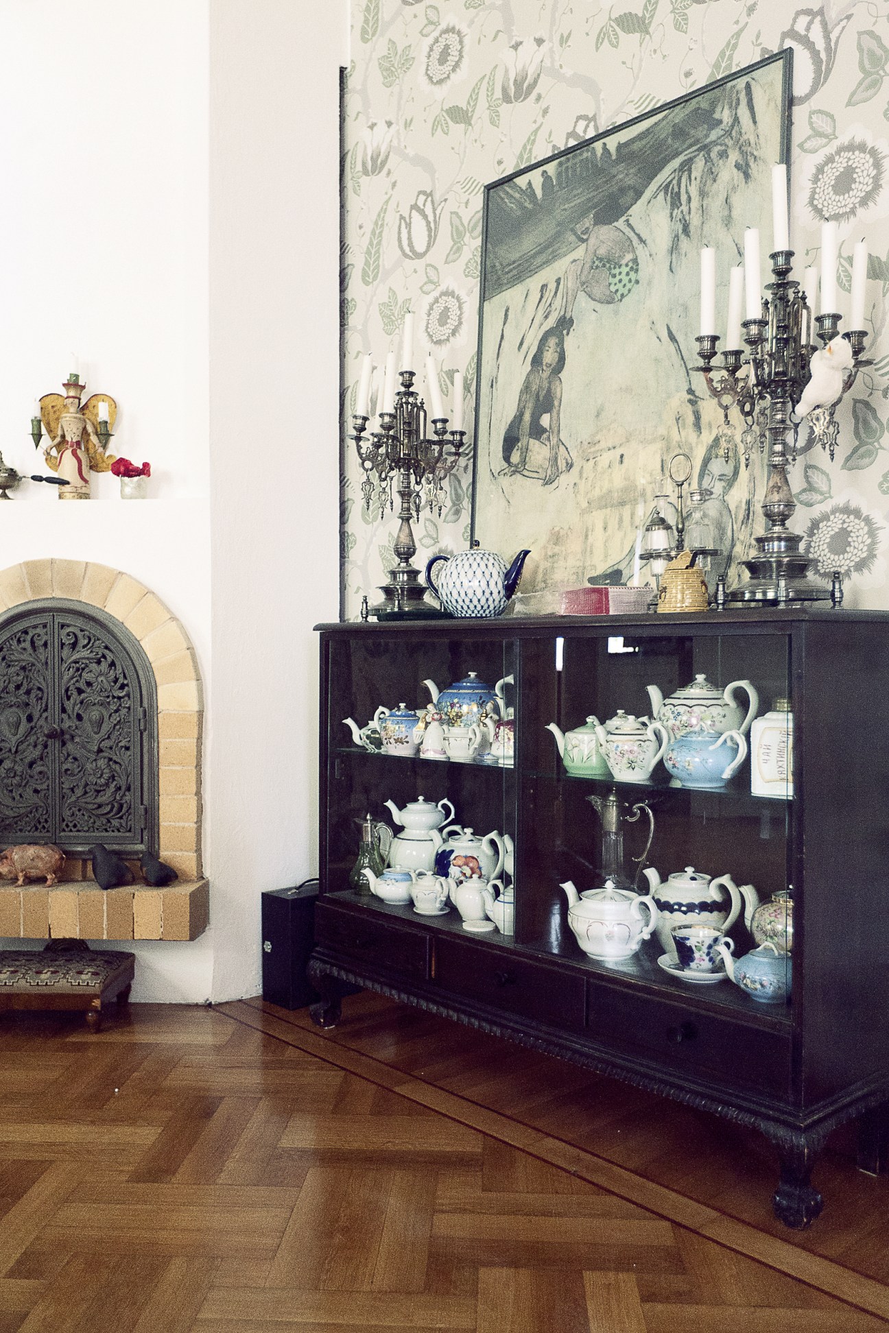 Display cabinet filled with decorative teapots and porcelain vessels against botanical wallpaper.