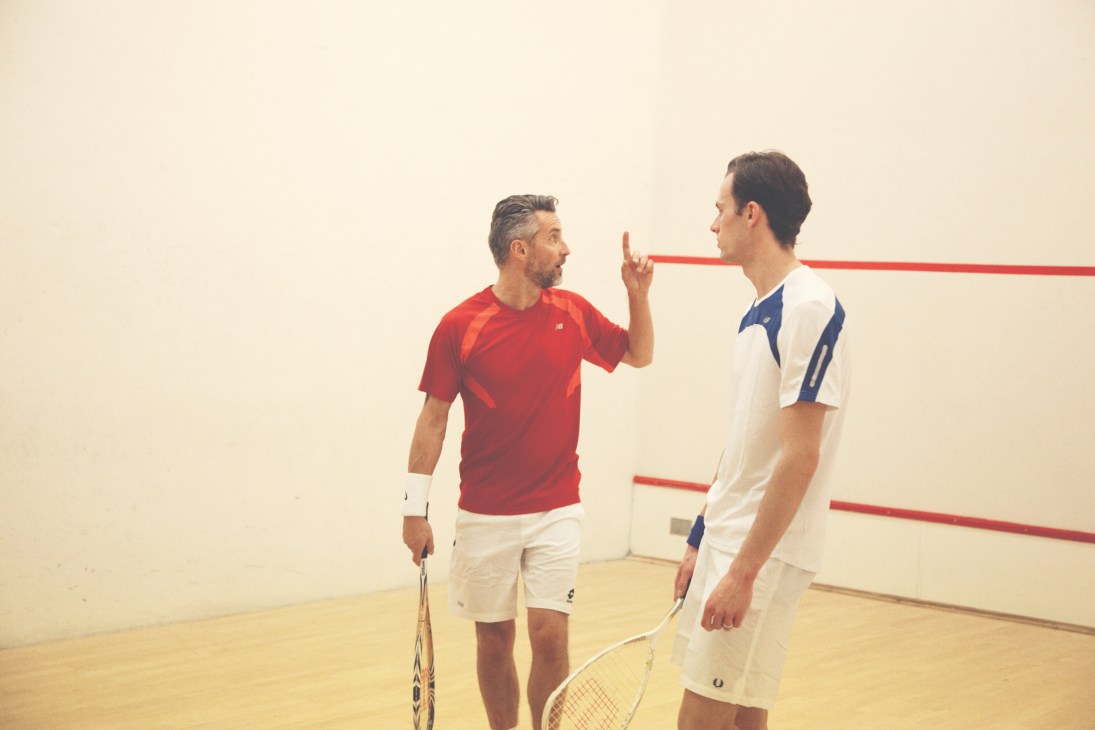 Two squash players facing each other on court, one in red New Balance shirt and the other in white and blue Wilson outfit.