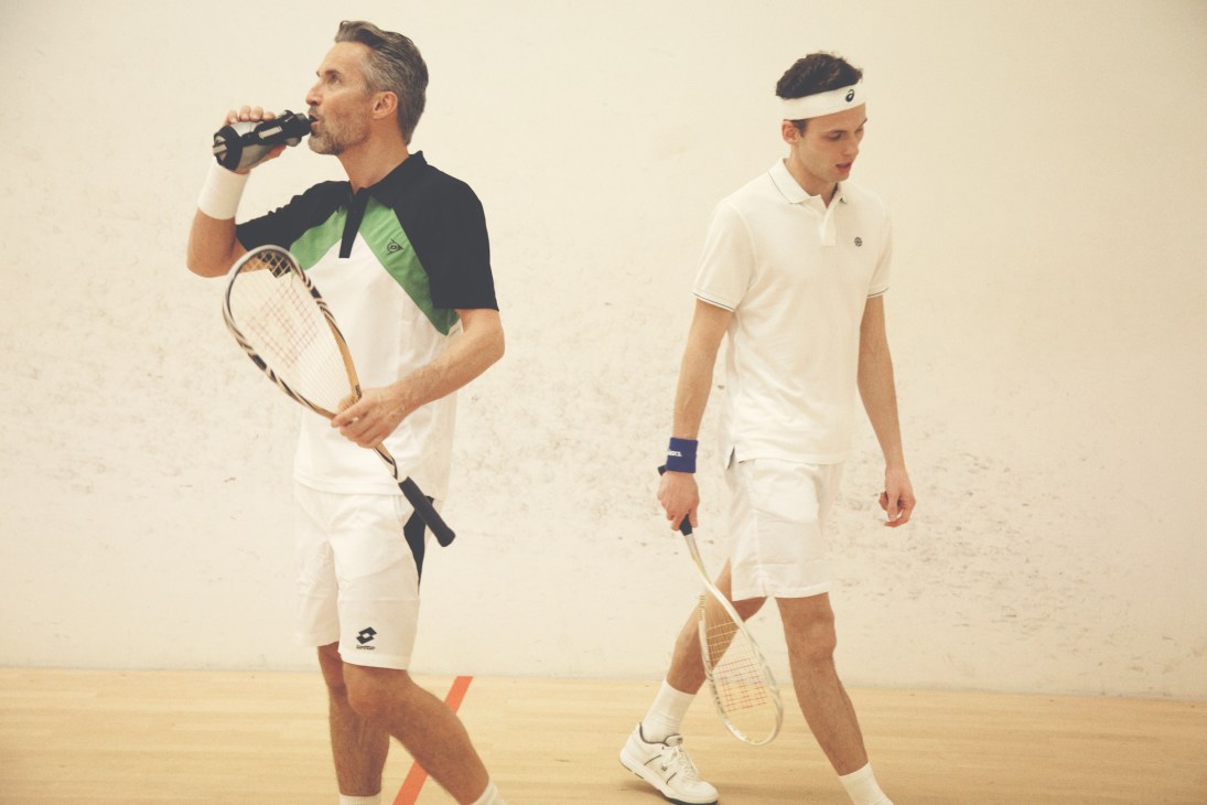 Two men in tennis wear on an indoor court; one drinks from a bottle while holding a racquet, the other stands nearby with a racquet.