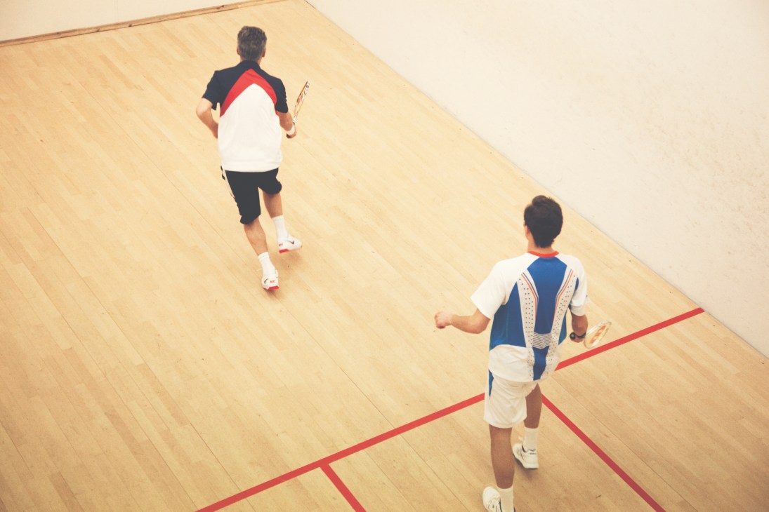 Two men playing squash on an indoor court with wooden floors and red boundary lines.