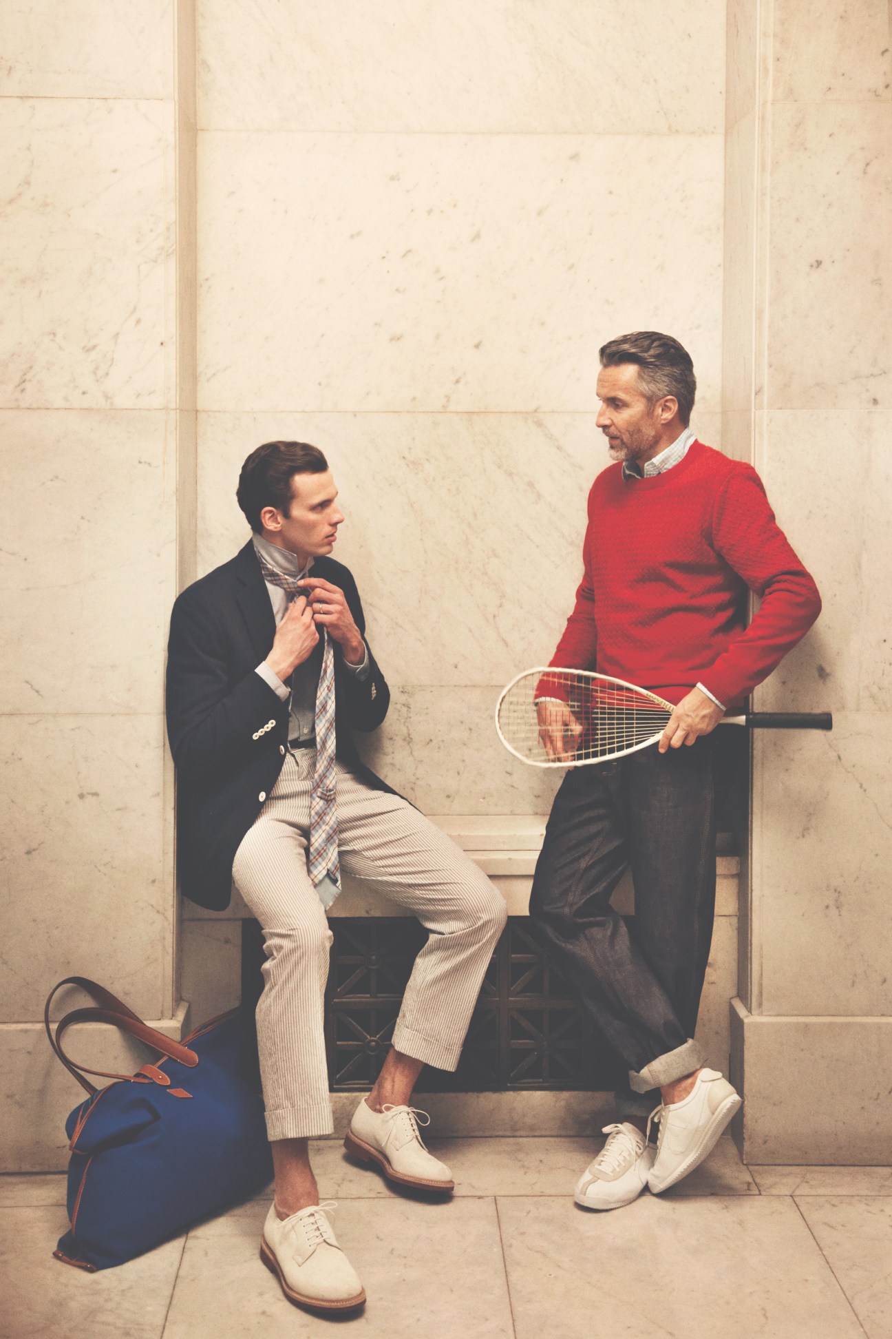 Two men in smart casual wear sitting on a bench with a tennis racquet, one in a red jumper and one in a black blazer.