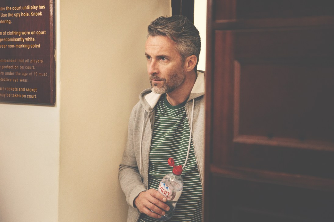 Man wearing Sunspel hooded top and Oliver Spencer t-shirt holds water bottle inside a clubhouse