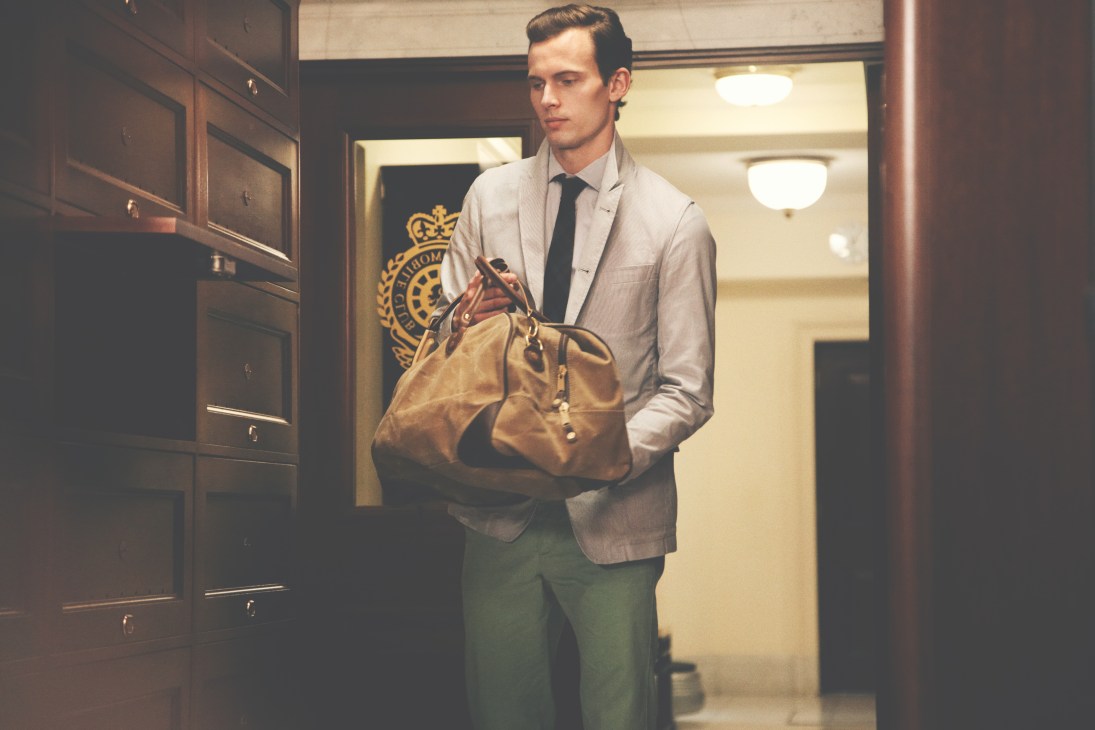 Man in Hackett shirt and Oliver Spencer jacket holding Woolrich bag at tennis club entrance.