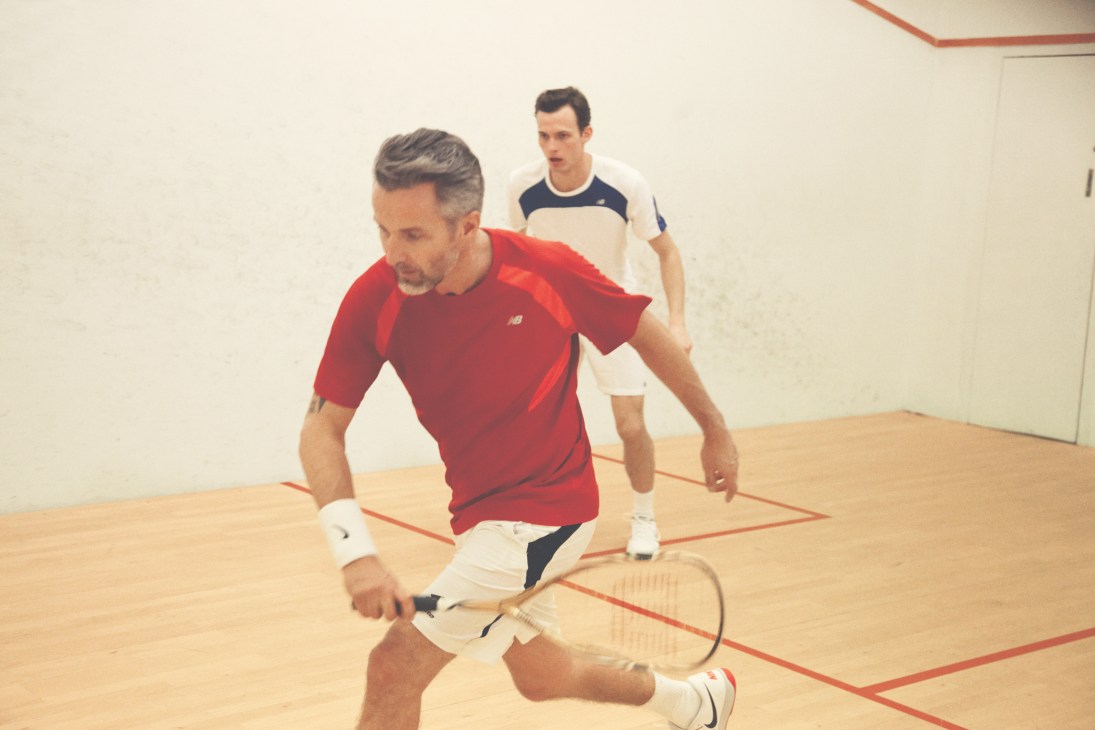 Two men playing squash on an indoor court, one in red New Balance shirt and the other in cream and navy striped shirt.