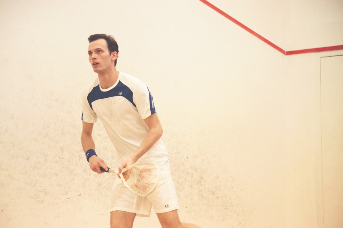Man in cream and navy tennis outfit holding Wilson racquet on indoor court with red trim.