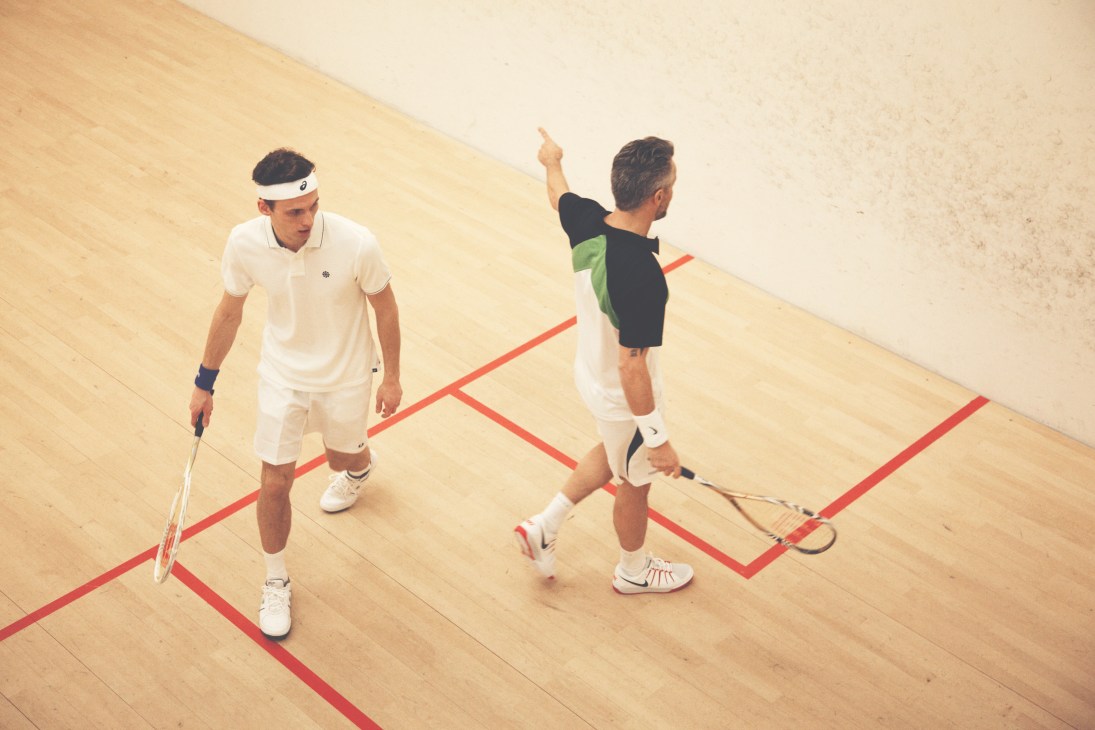Two men playing squash on a wooden court with red boundary lines, one wearing white and one wearing black and green.