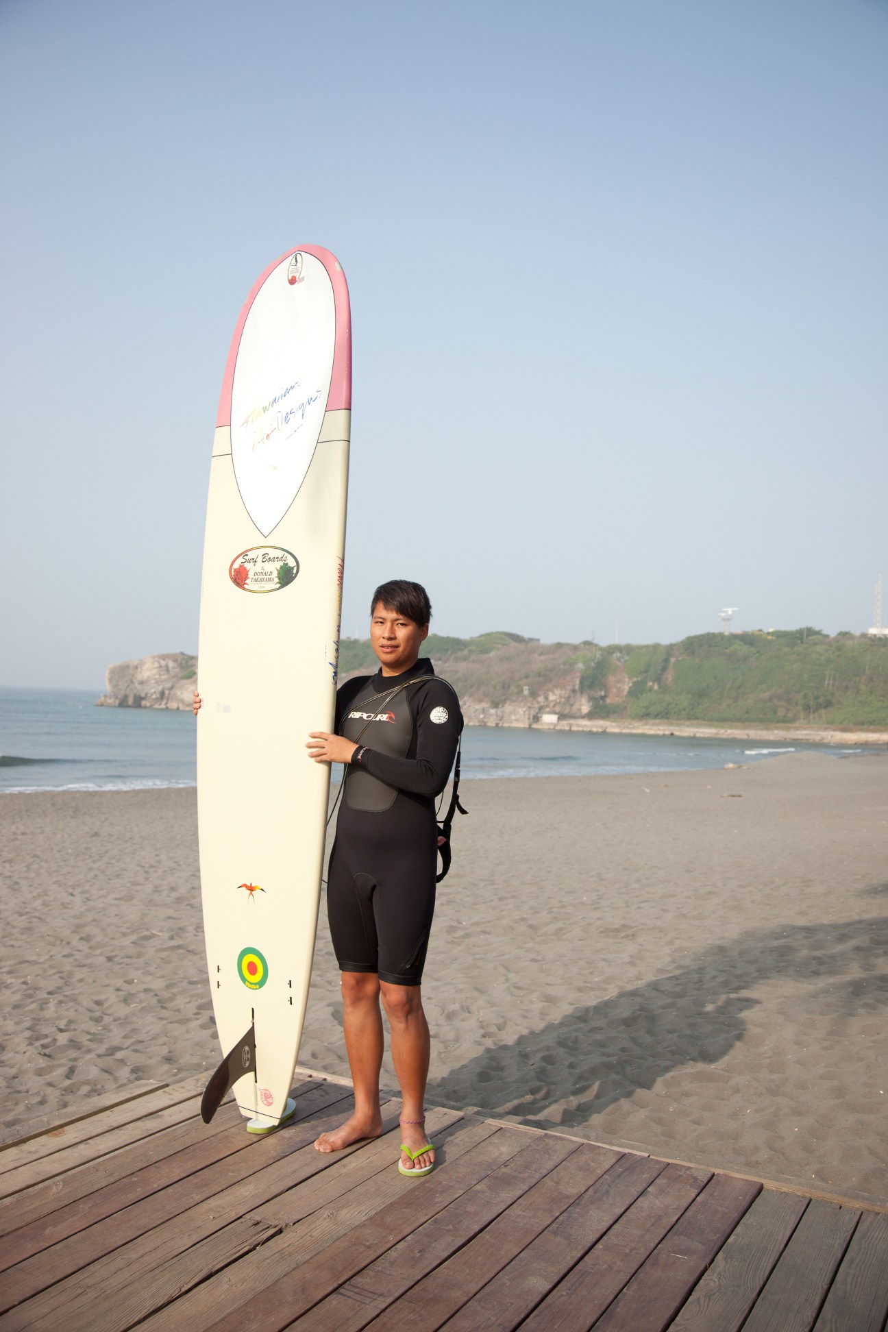 Young surfer in black wetsuit holding surfboard on Kaohsiung beach with rocky coastline in background.