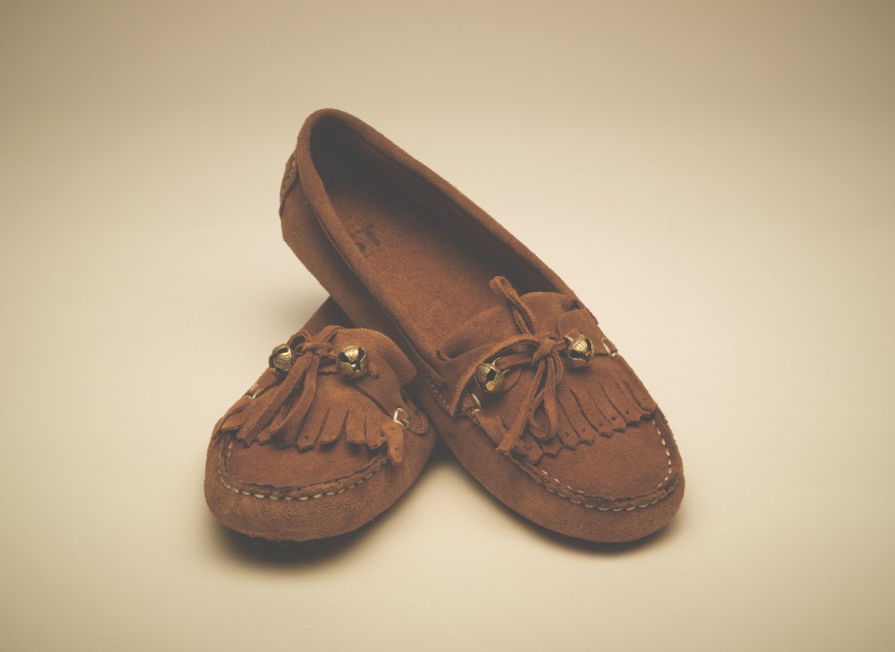 Brown suede moccasins with decorative fringe and bells on a neutral background.