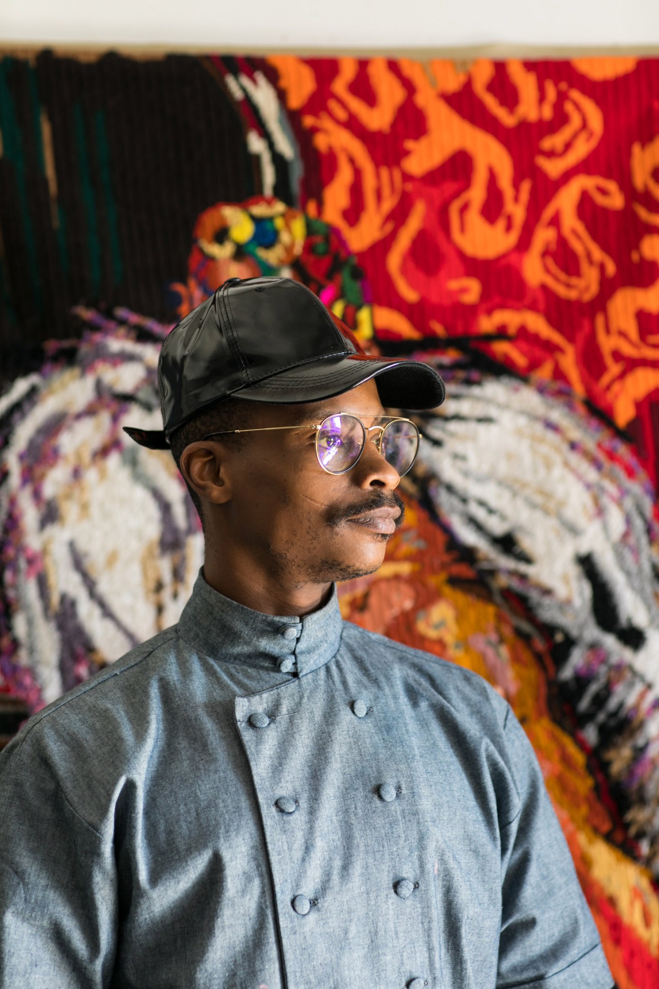 Athi-Patra Ruga wearing a black cap and round glasses stands in front of colorful patterned artwork.