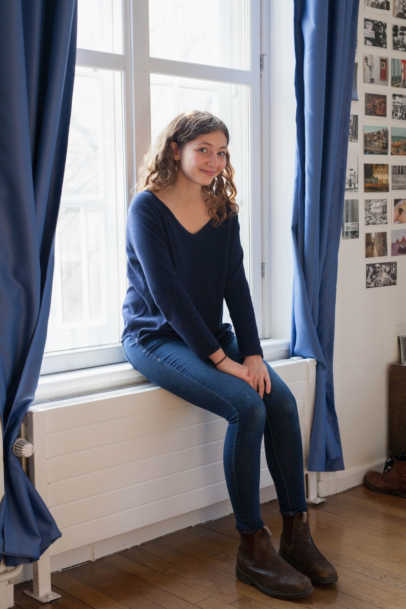 Woman sitting on a windowsill with blue curtains, smiling at the camera in a dormitory room decorated with photographs.