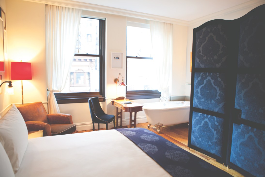 NoMad hotel bedroom with Jacques Garcia interiors, featuring a white bed, orange armchair, and freestanding bathtub by windows.
