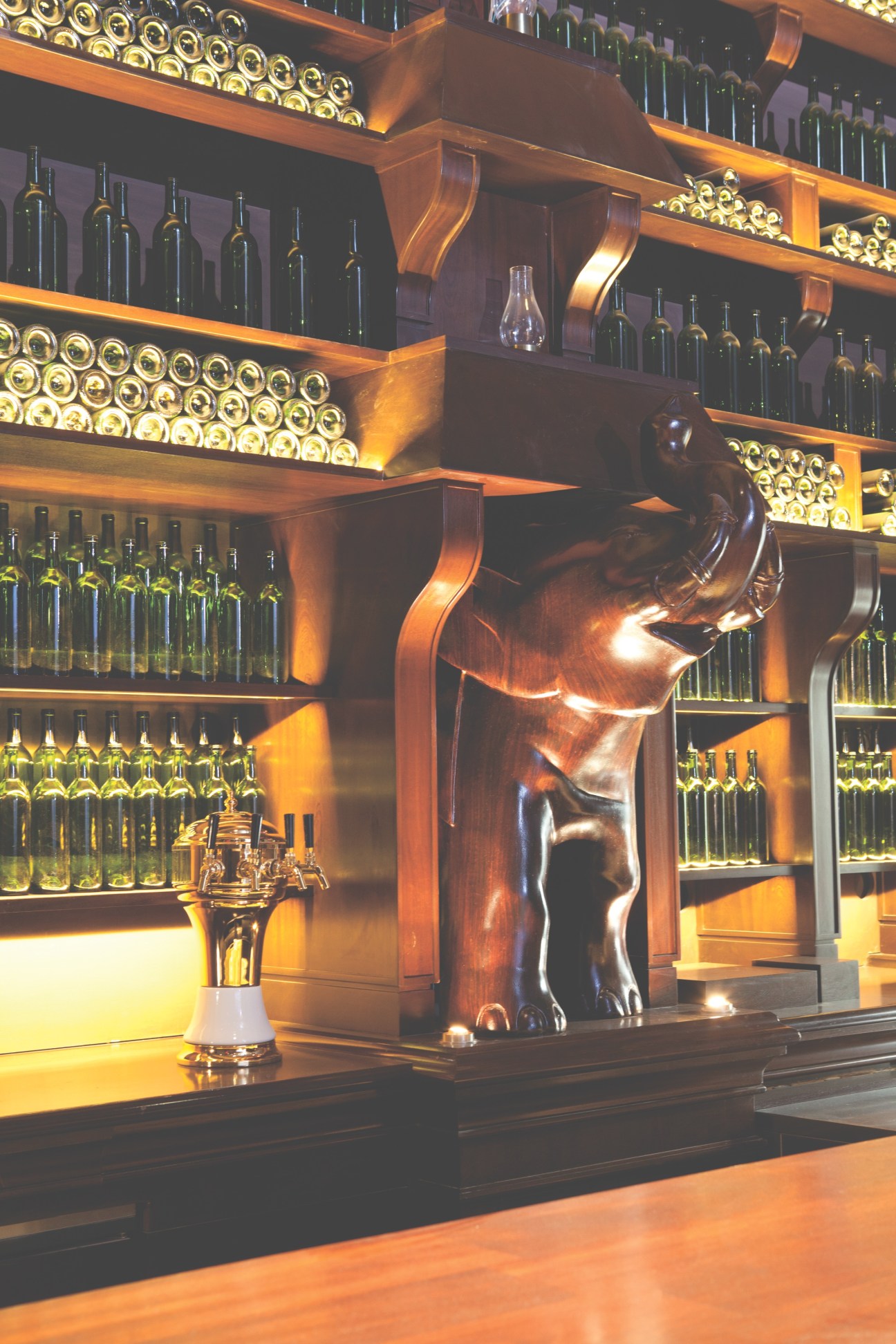 NoMad hotel's ground floor bar featuring illuminated wine shelves, ornate wooden sculpture, and warm ambient lighting.