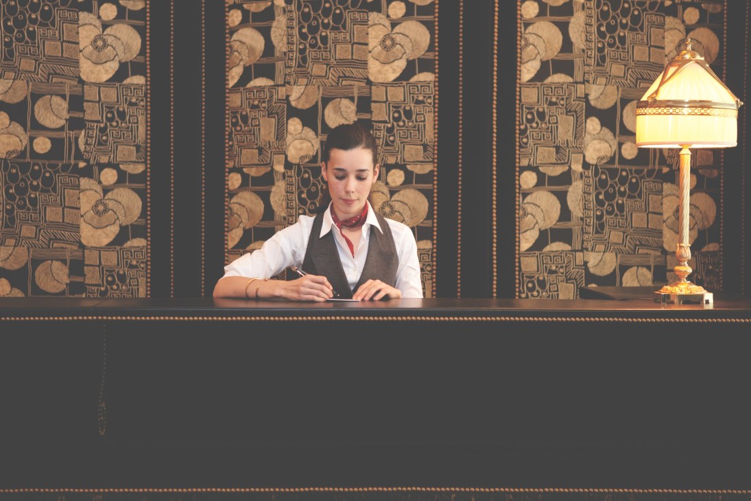 Hotel receptionist at check-in desk with patterned wallpaper backdrop and brass lamp.