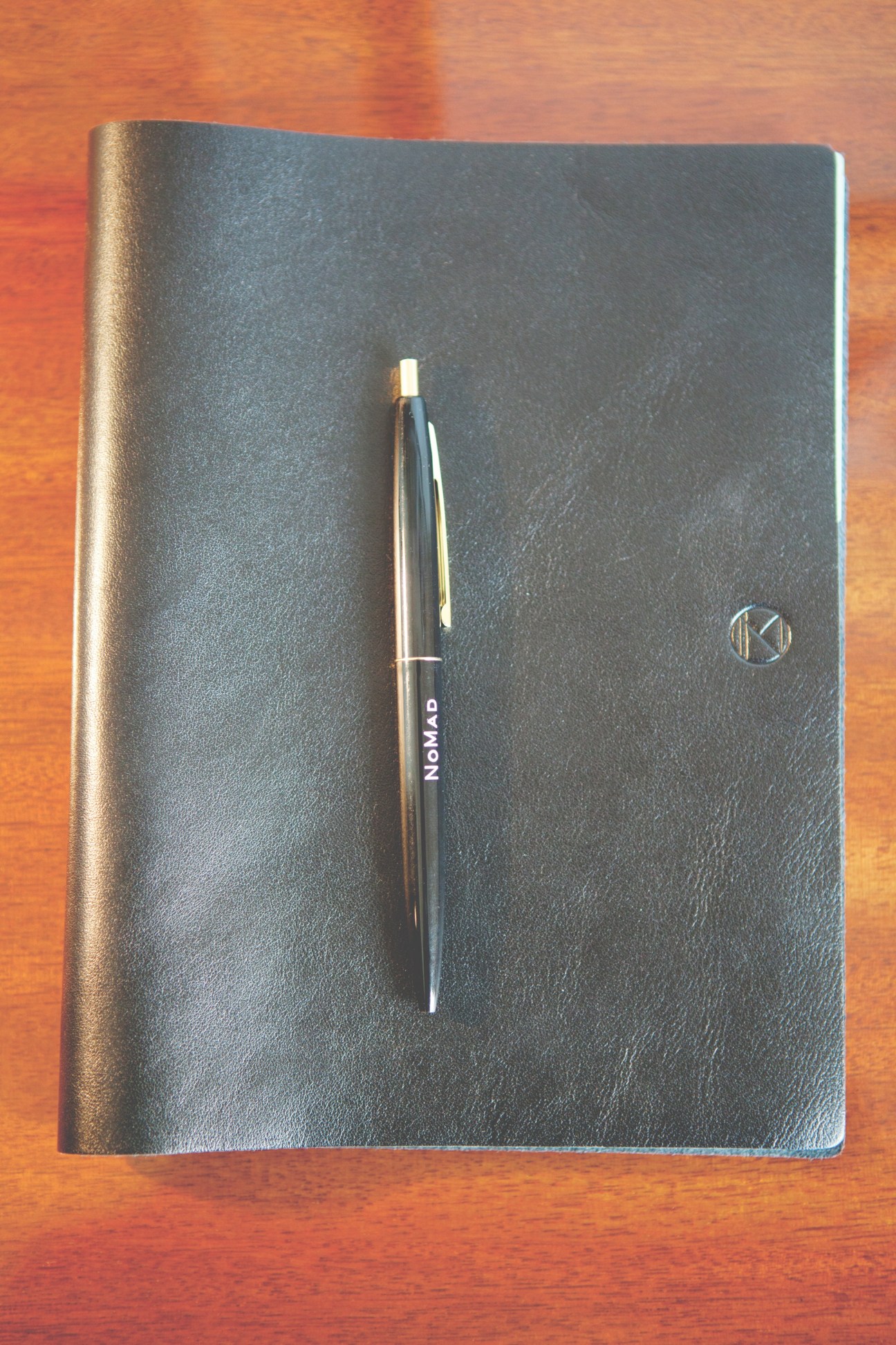 Dark leather notebook with gold pen and WANT Les Essentiels de la Vie logo on wooden surface.