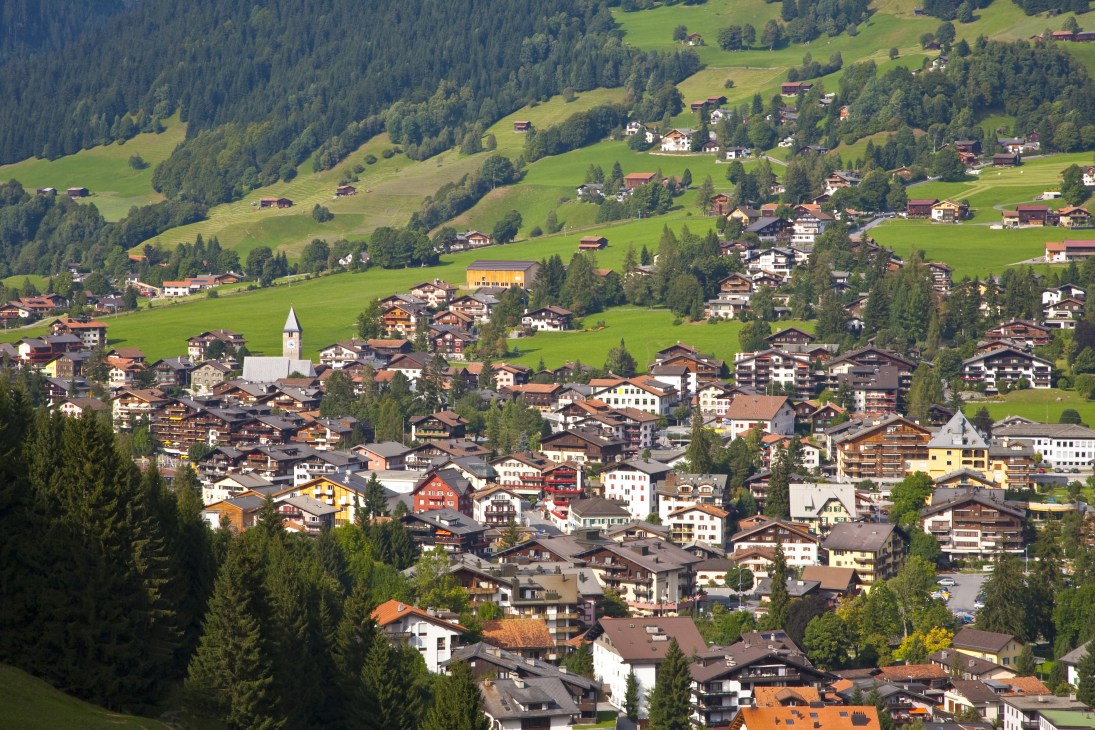 Aerial view of a dense Alpine village with chalets and buildings nestled in a green mountain valley surrounded by forests.