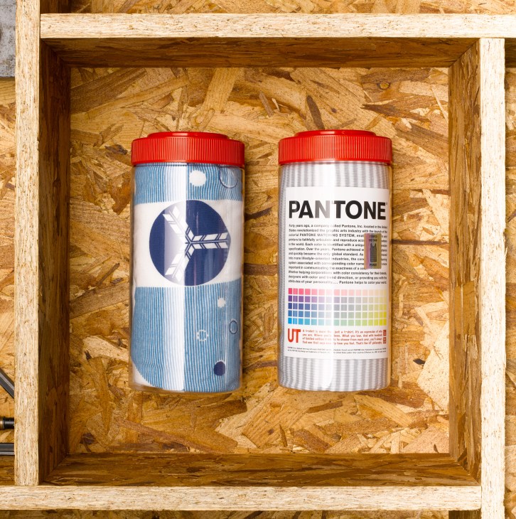Two cylindrical containers with red caps in a wooden box display, one with blue geometric design and one showing Pantone color matching system.