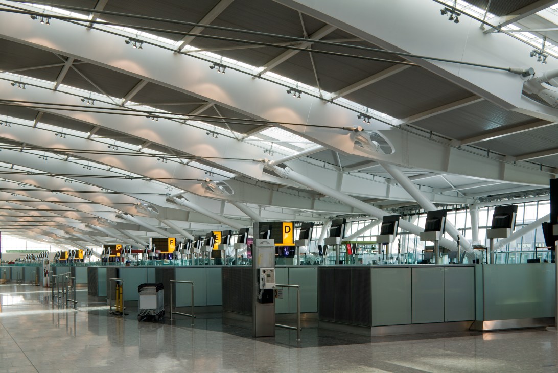 Heathrow Terminal 5 check-in area with white structural girders, glass panels, and illuminated gate signs.