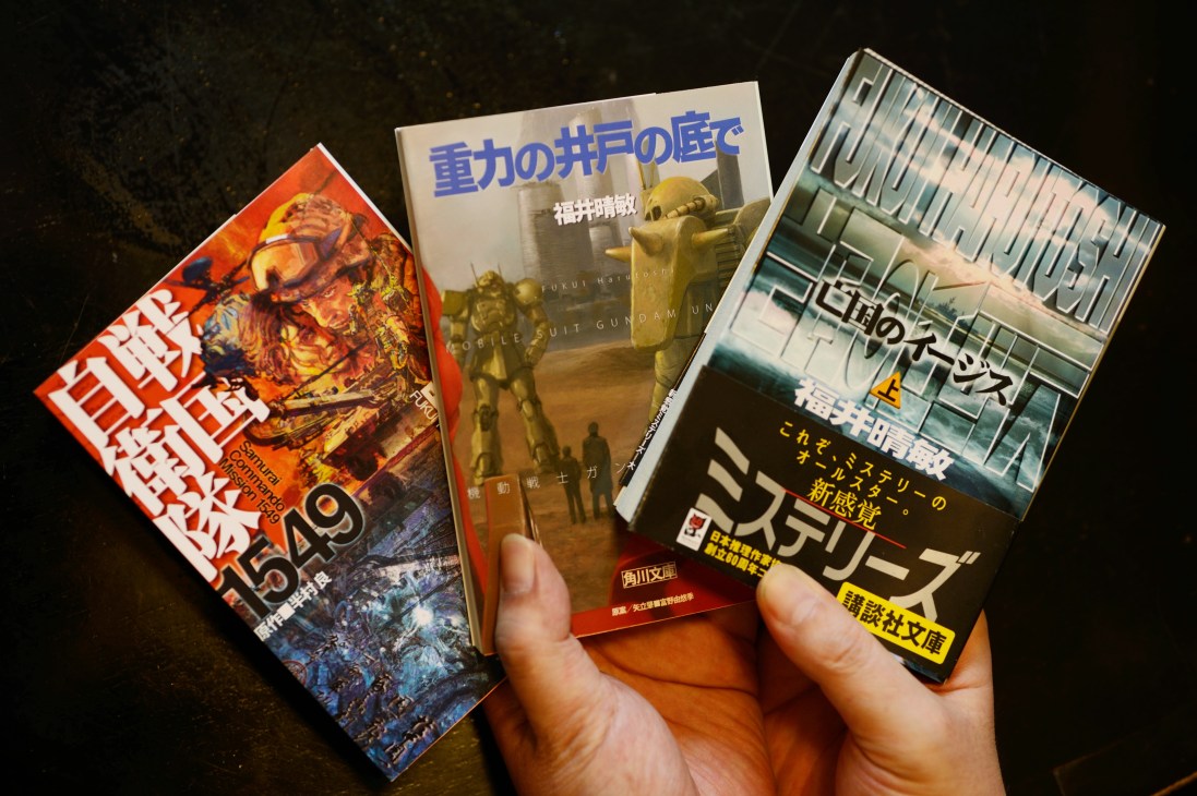 A hand holding Japanese novels and DVDs about military themes, including works by Harutoshi Fukui.