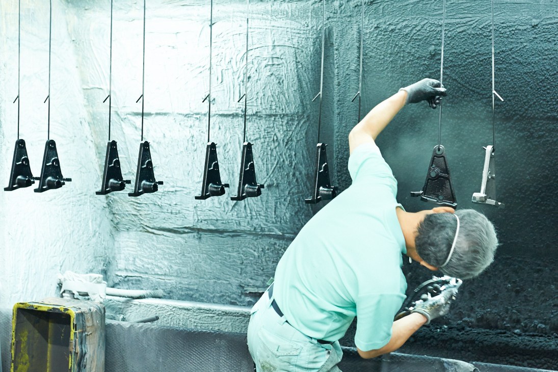 Worker inspecting metal chair components hanging from strings in Takara Belmont foundry factory.
