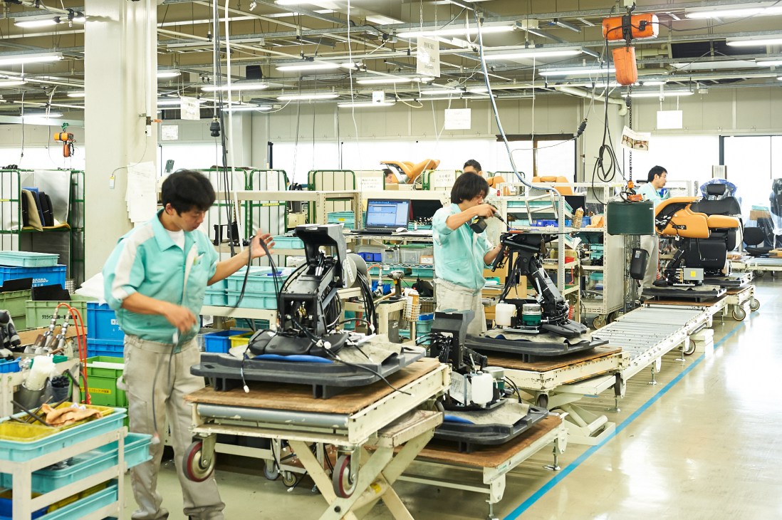 Workers in turquoise uniforms assemble barber and salon chairs at Takara Belmont factory workstations.