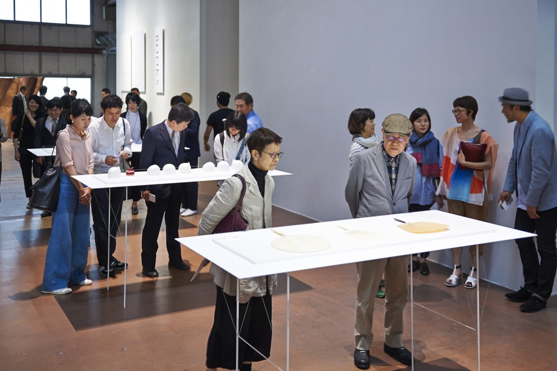 Visitors explore the Takeo Paper Show exhibition in a warehouse gallery, examining white paper displays on tables.