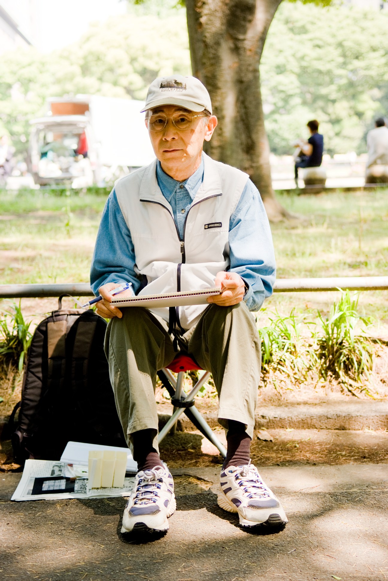 Taku Ito, 70, sits outdoors holding a sketchpad while painting, wearing a white cap and vest.