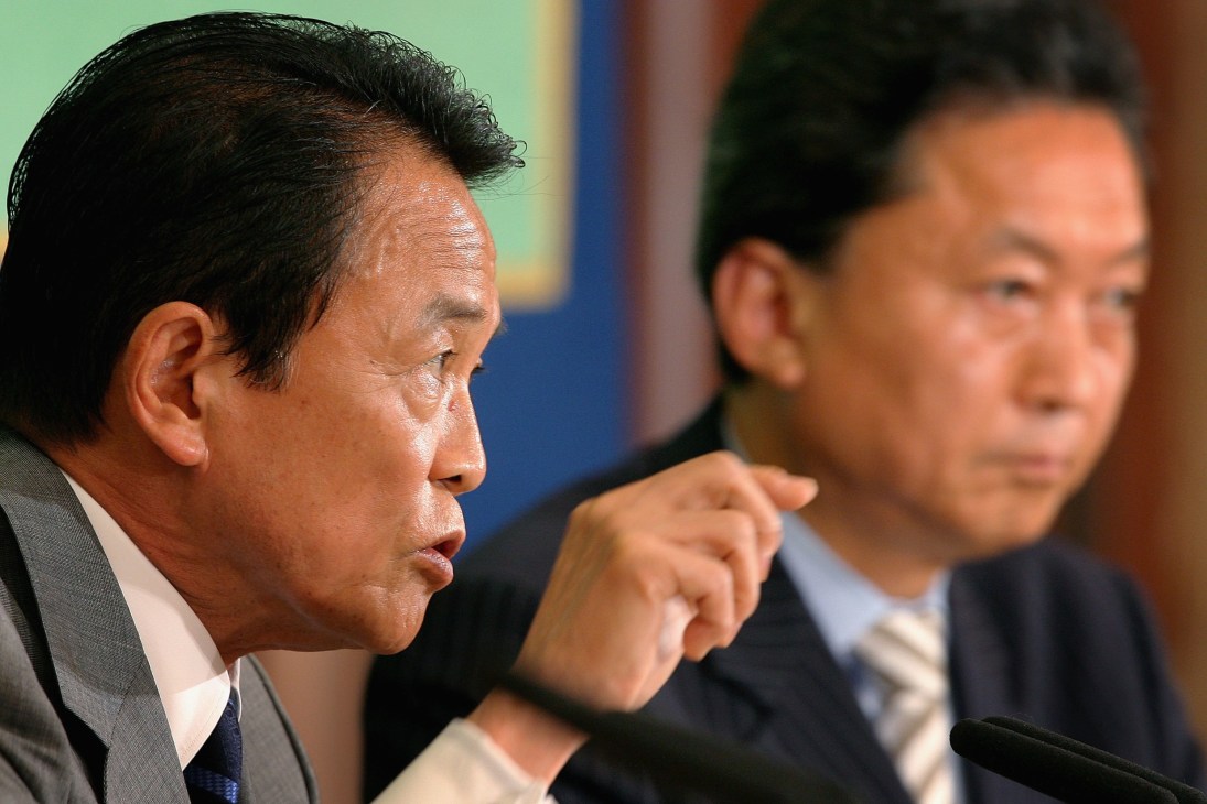 Taro Aso speaking at a press conference with another official seated beside him.