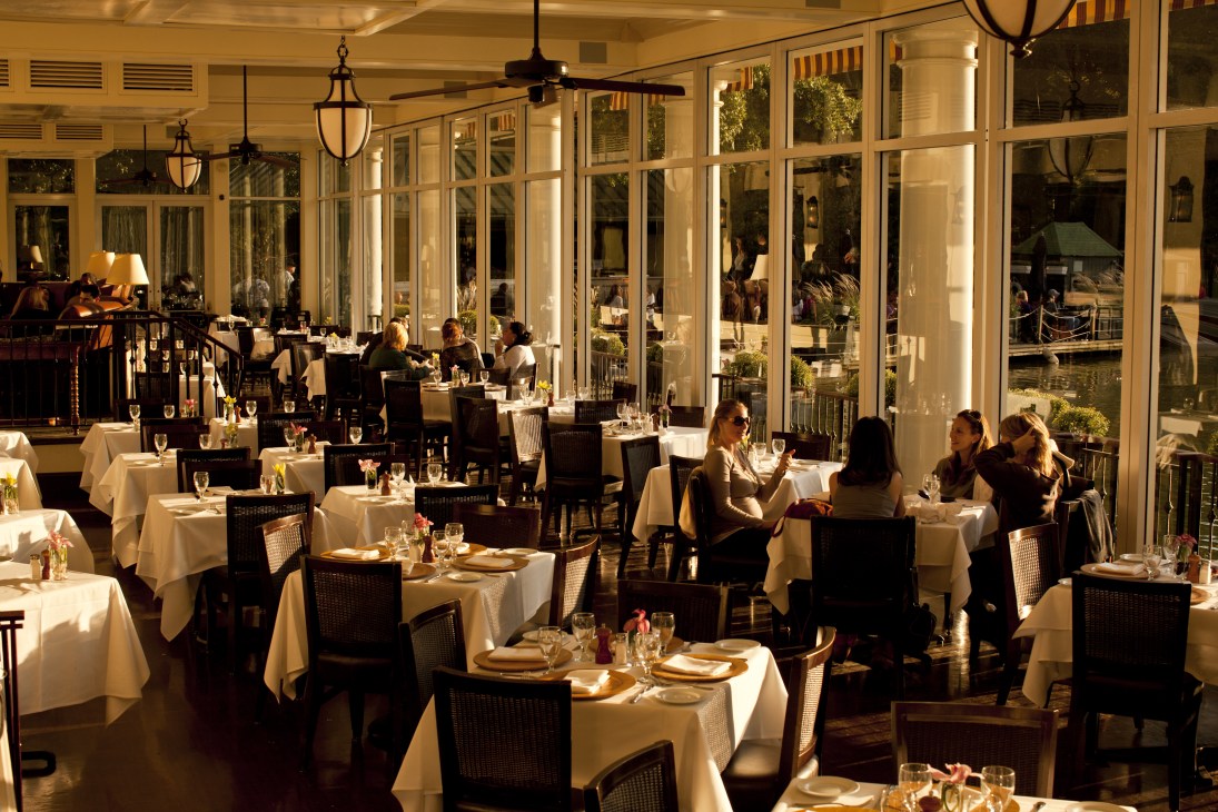 The Central Park Boathouse dining room with white tablecloth tables, black chairs, large windows overlooking the lake, and diners seated throughout.