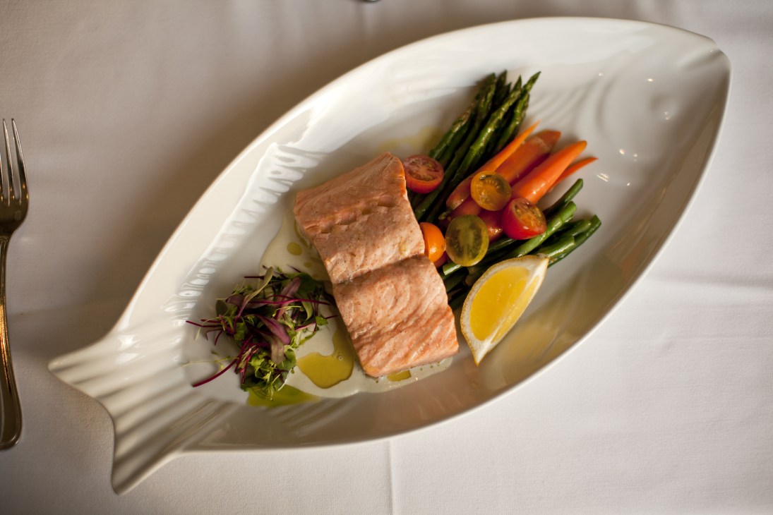 Poached salmon fillet with asparagus, carrots, tomatoes and lemon on a white fish-shaped plate.
