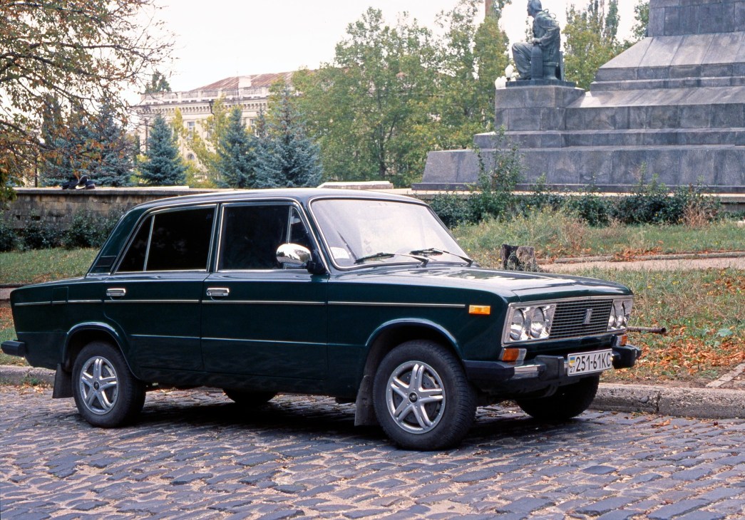 Dark blue Lada 2105 sedan parked on cobblestone street with Eastern European architecture and monument in background.