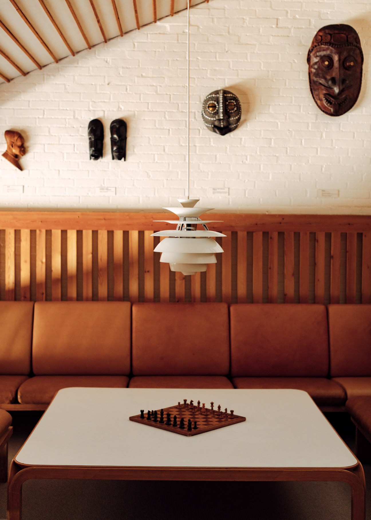 Mid-century modern interior with cognac leather sofa, white pendant lamp, wooden wall paneling, and African masks and artifacts on white brick wall above.
