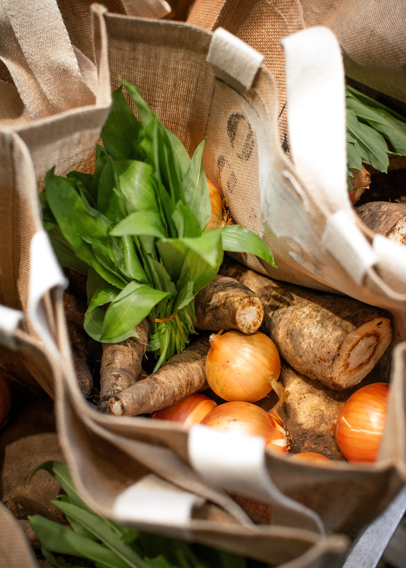 Natural jute bag filled with organic vegetables including leafy greens, onions, and root vegetables.