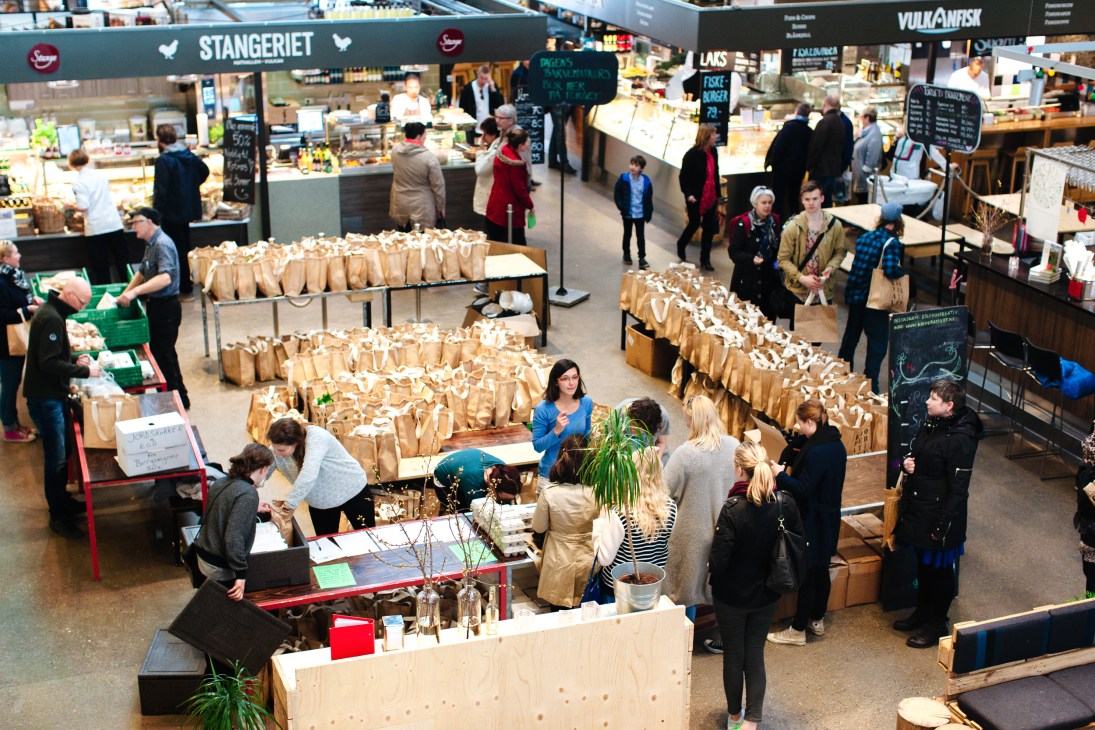 Overhead view of Kooperativet's pick-up point at Mathallen food market in Oslo, with paper bags of organic produce arranged on tables and customers shopping.