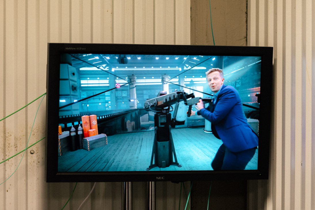Host Fridtjof Nilsen holds a rifle prop while standing in a futuristic virtual environment displayed on a monitor.