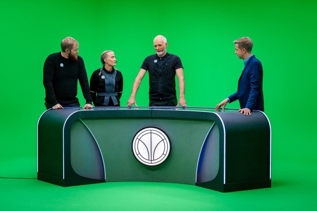 Four people stand around a futuristic game show control panel with a green screen background.