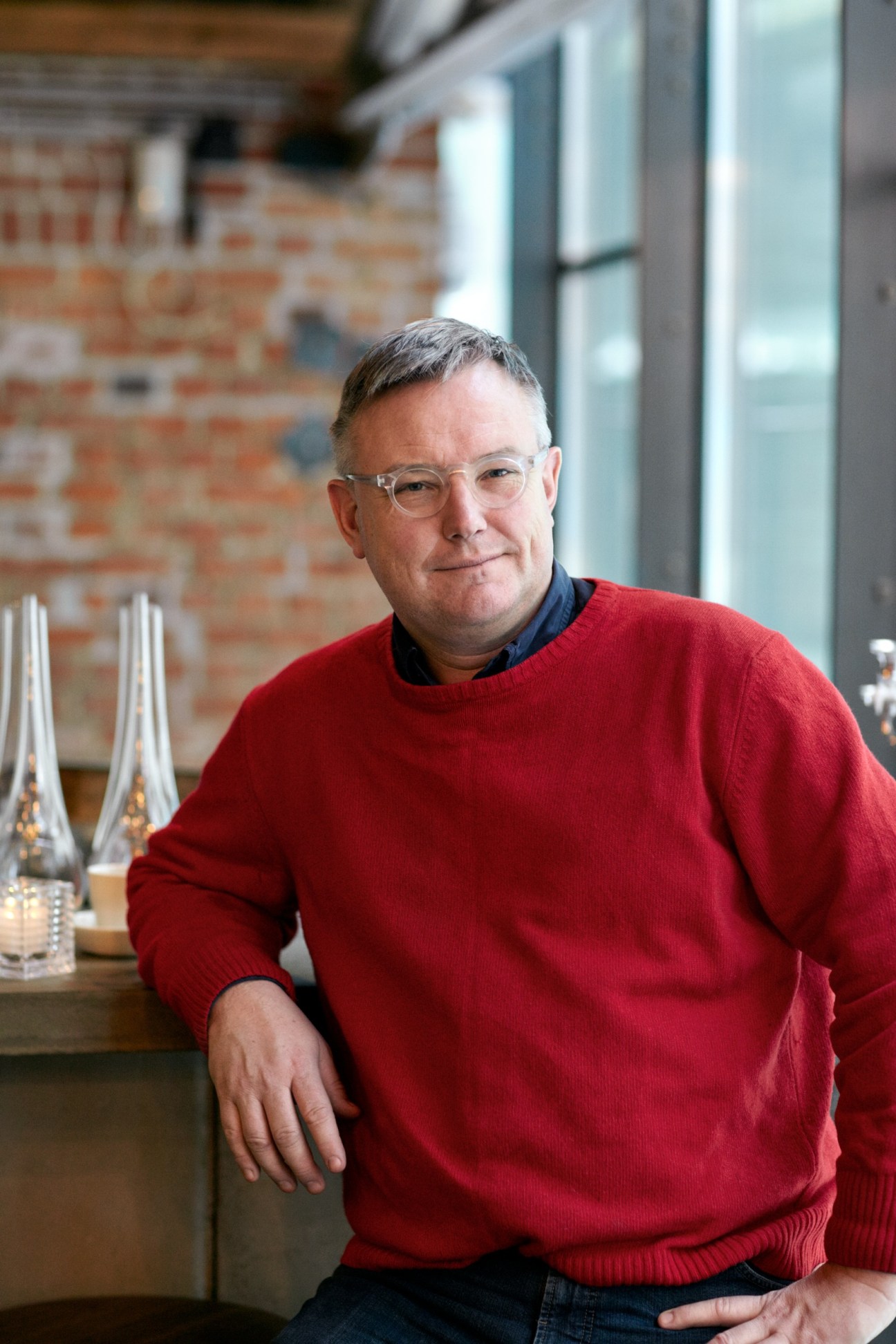Man wearing glasses and red sweater sits in front of exposed brick wall at Mathallen food hall.