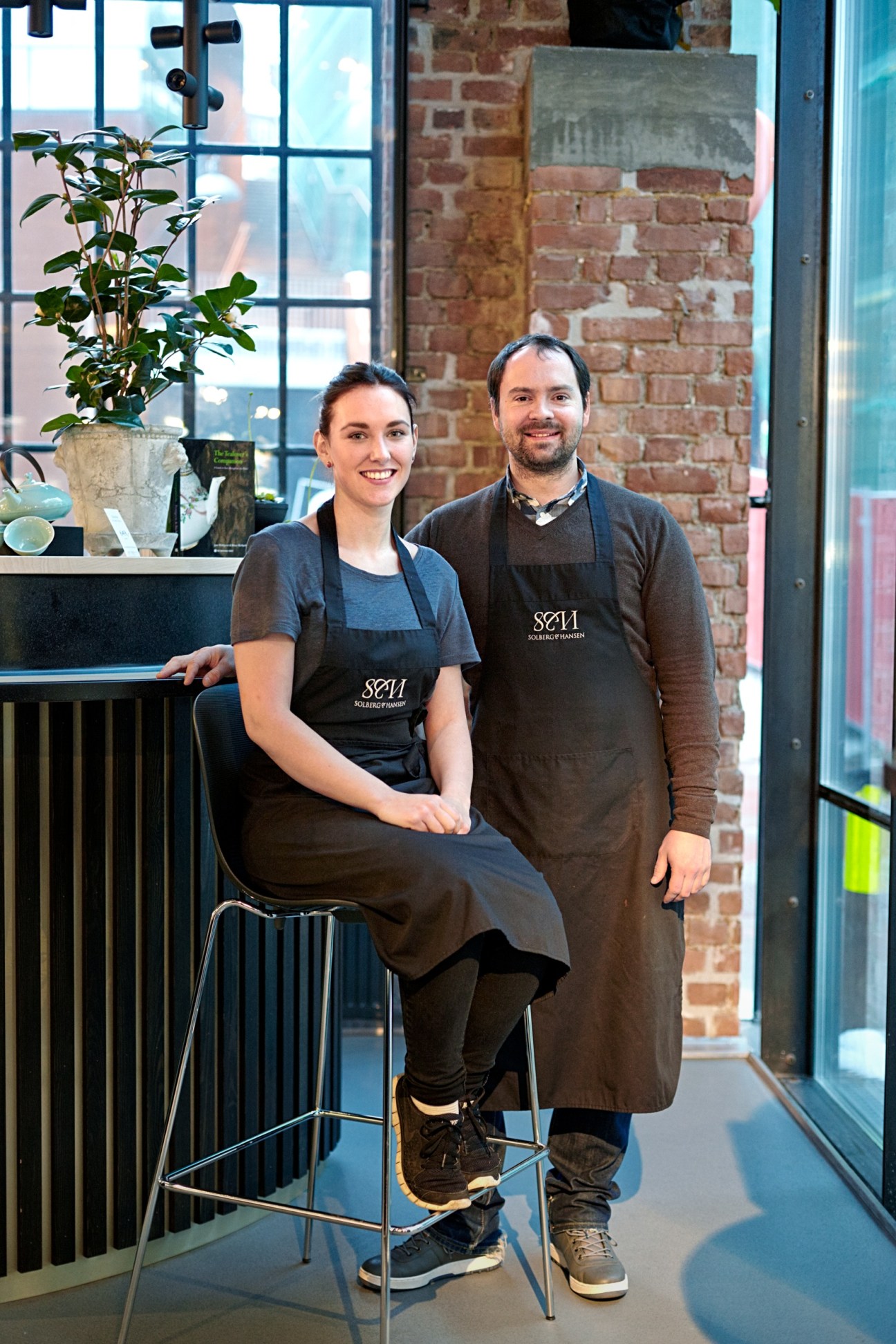 Two staff members wearing Solberg & Hansen aprons stand together inside the food hall with exposed brick wall behind them.