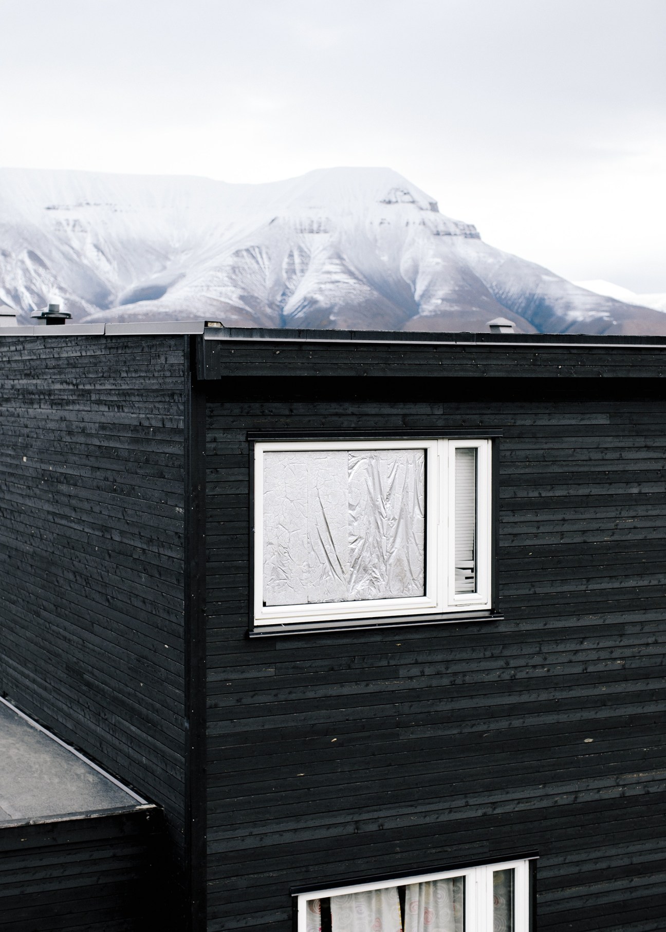 Black wooden house with tin foil-covered window blocking midnight sun, snow-covered mountain in background.