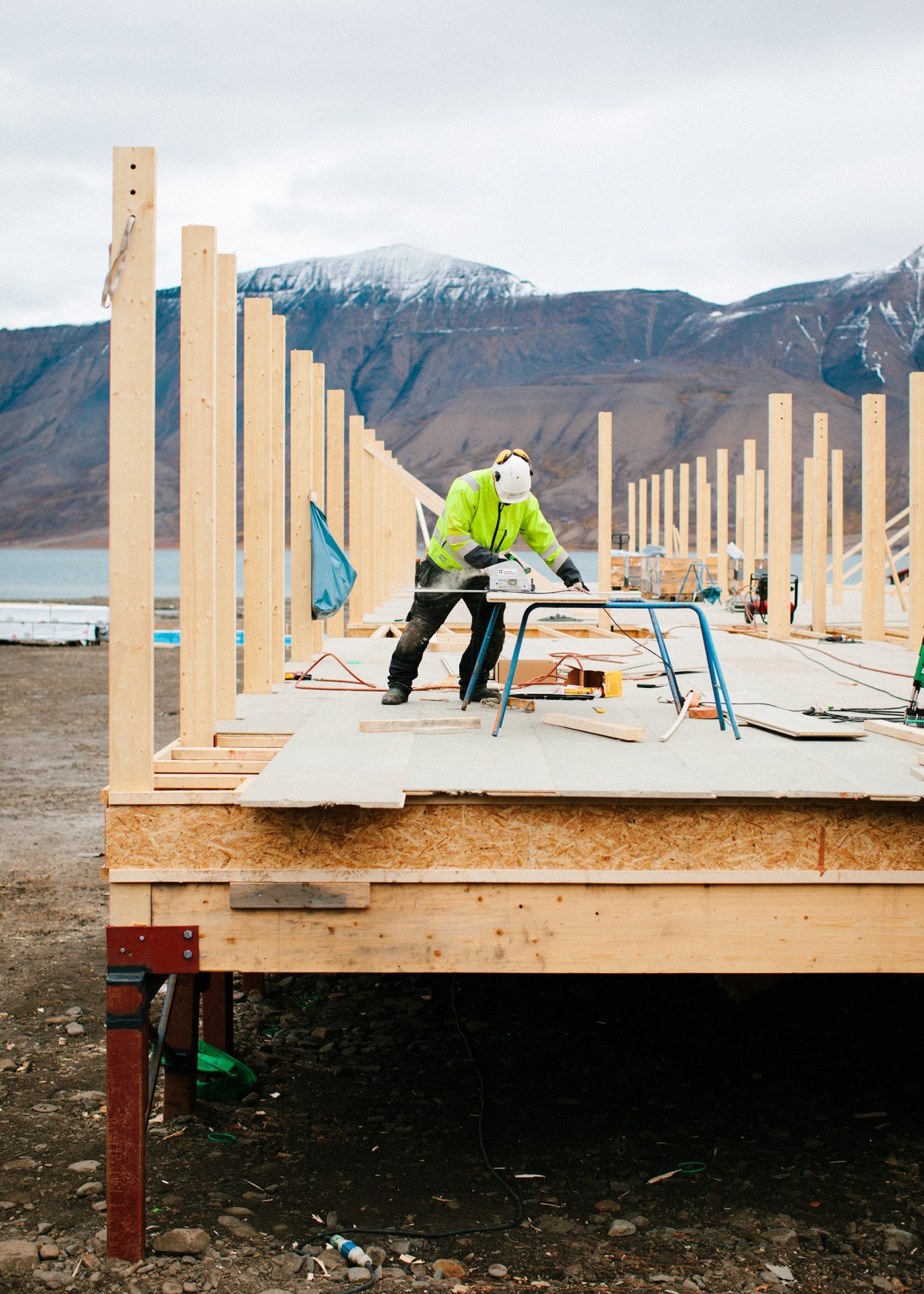 Worker in high-visibility jacket constructing raised wooden foundation for student housing in Svalbard, with snow-capped mountains in background.