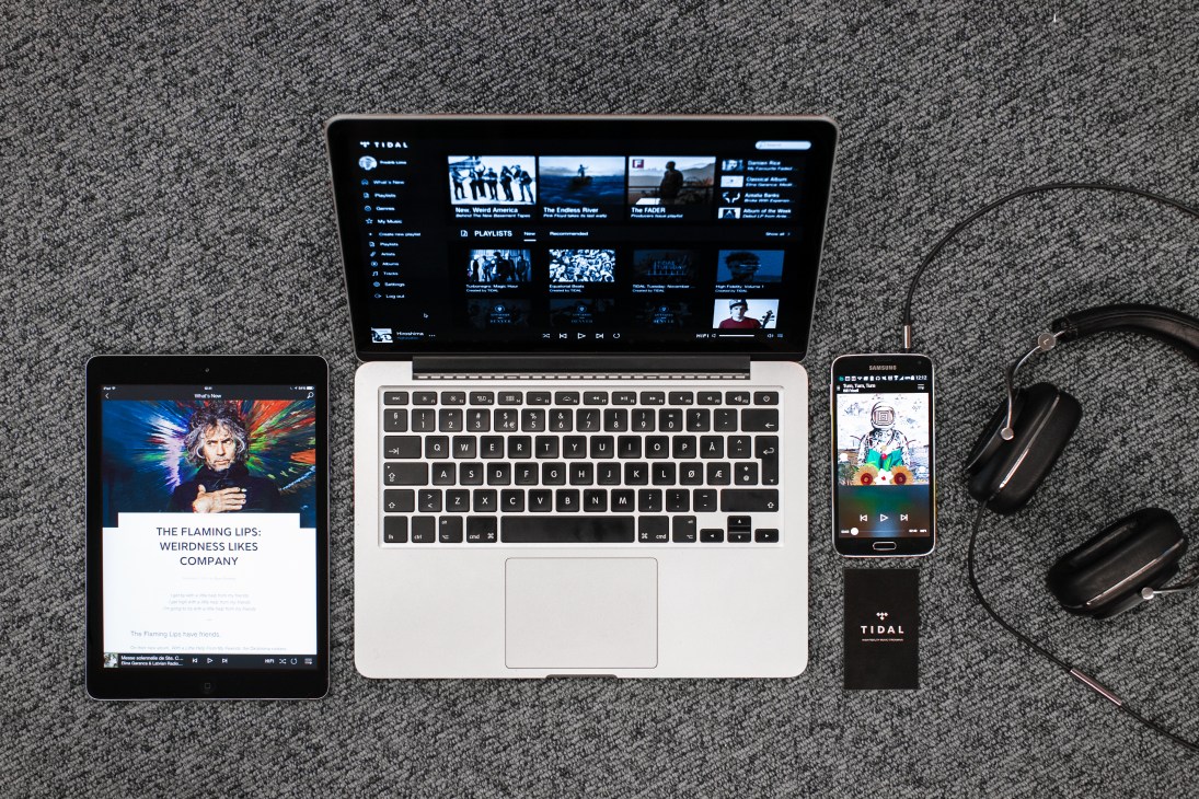 Tidal music streaming service displayed on laptop, tablet, smartphone and portable device with headphones on textured gray surface.