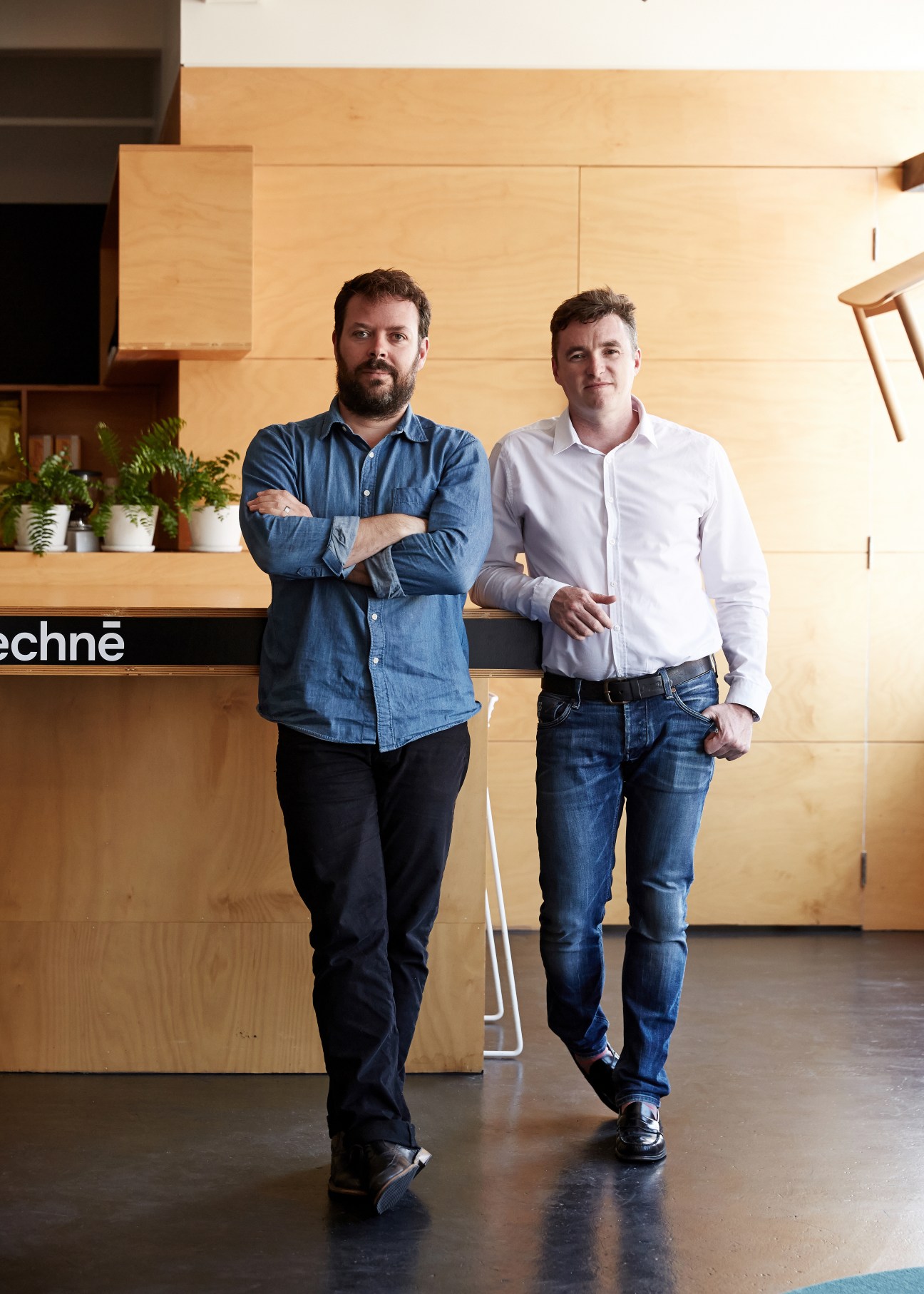 Two men standing in a wooden interior studio space with minimalist furniture and plants visible behind them.