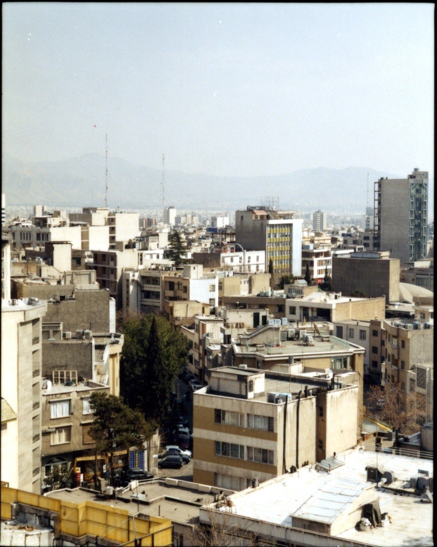 Aerial view of downtown Tehran showing dense residential buildings under hazy skies with mountains in the distance.