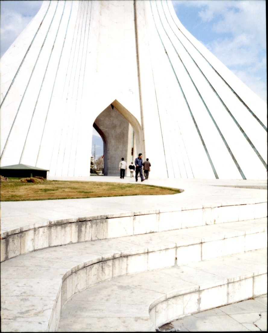 The Azadi Tower in Tehran with its distinctive white arched structure and people walking through the central gateway.