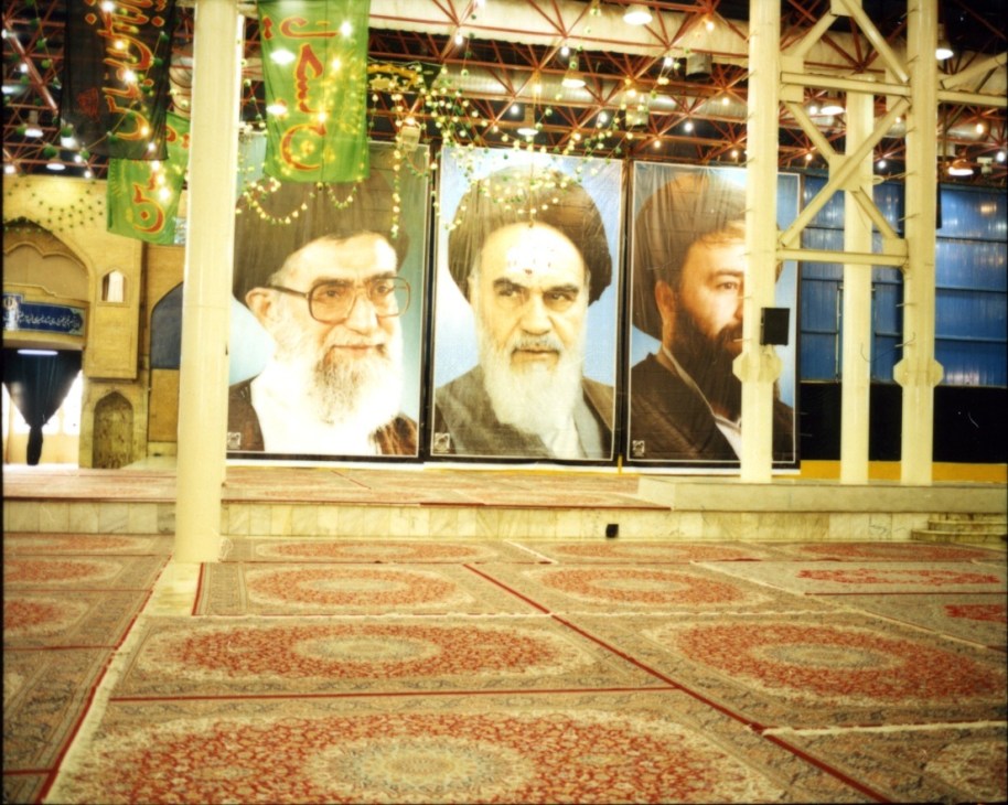 Interior of Iman Khomeini mosque with three large portrait posters of religious leaders displayed on the wall above an ornate patterned carpet.