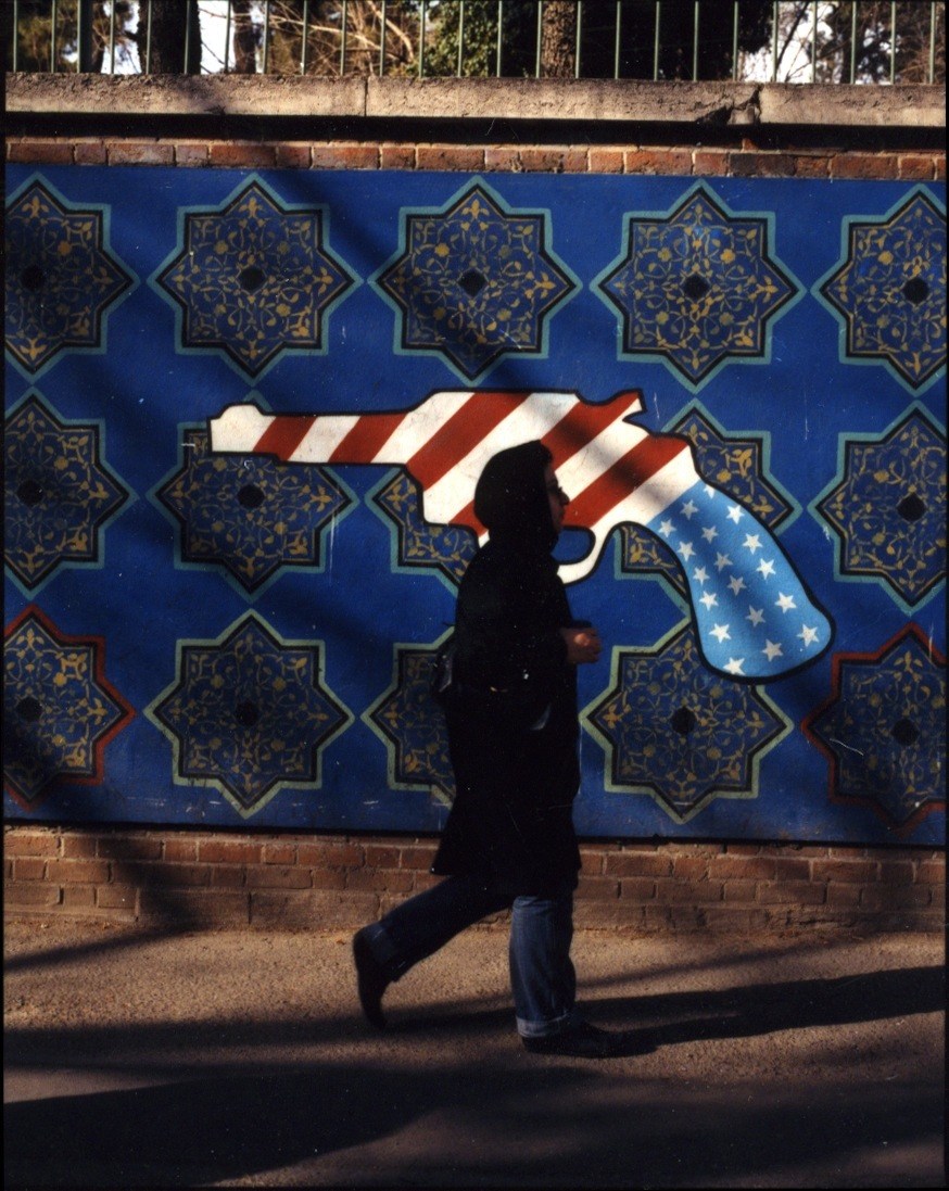 Silhouetted figure standing before anti-American mural with US flag imagery on blue Islamic tile background in Tehran